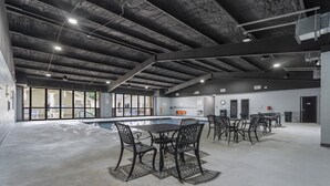 A spacious indoor area with a pool, tables, and chairs, featuring a concrete floor and exposed ceiling beams.