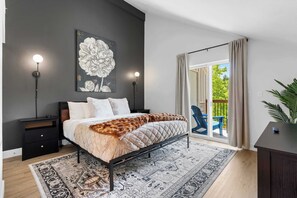 A bedroom with a bed, a nightstand, a wall-mounted lamp, a large floral painting, and a view of the outdoors through a sliding door. - Creekside Duo Near the Landing (Branson)