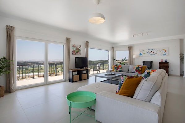 A spacious living room with a sofa, a coffee table, and a TV. Large windows offer a view of the outdoors.