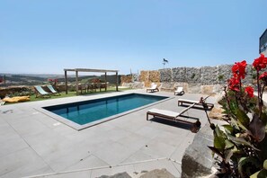 A rectangular swimming pool with lounge chairs and a pergola, surrounded by a stone wall and a view of the countryside.