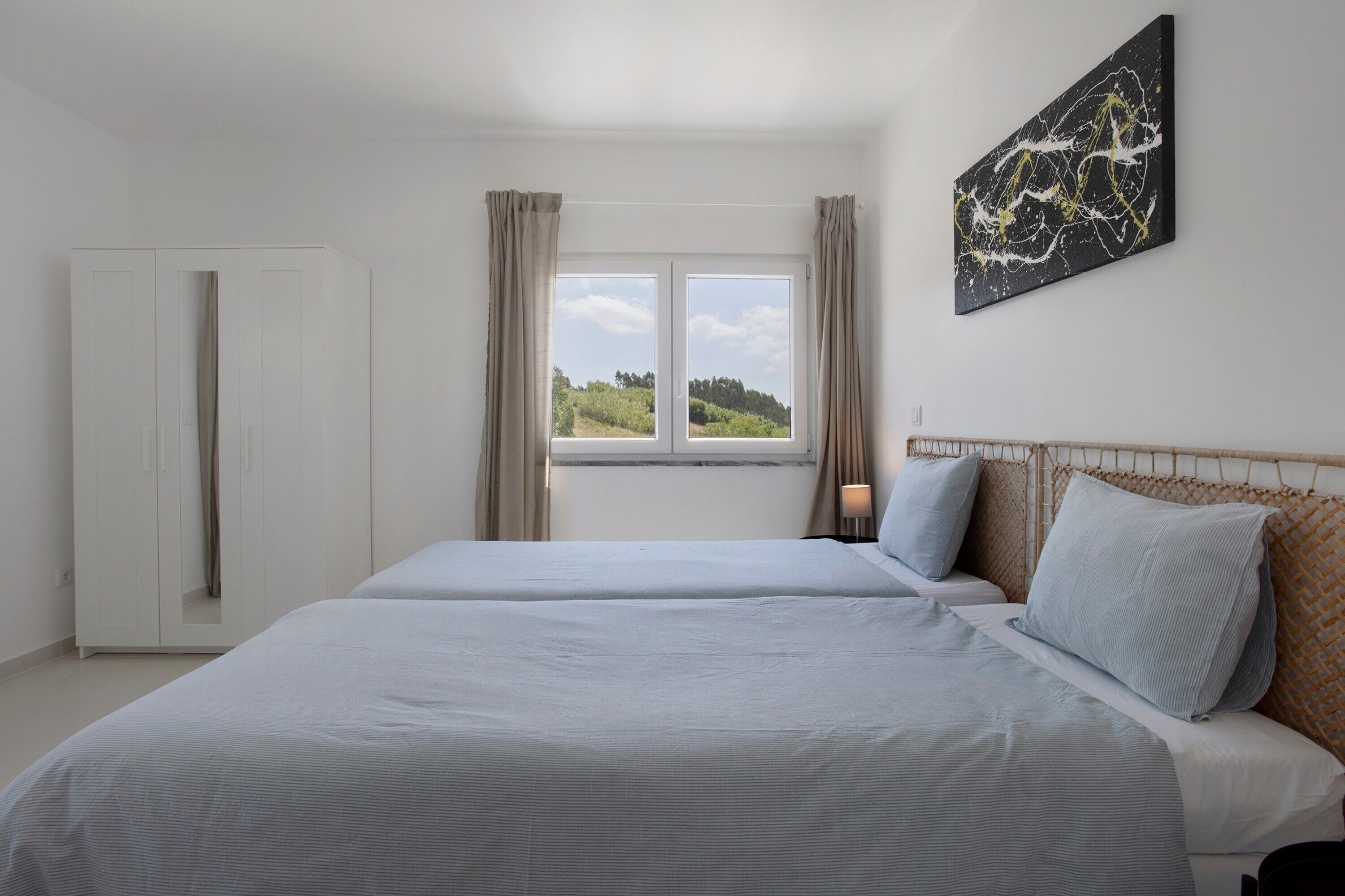 A bedroom with two beds, a window with curtains, a wardrobe, and a wall-mounted artwork.