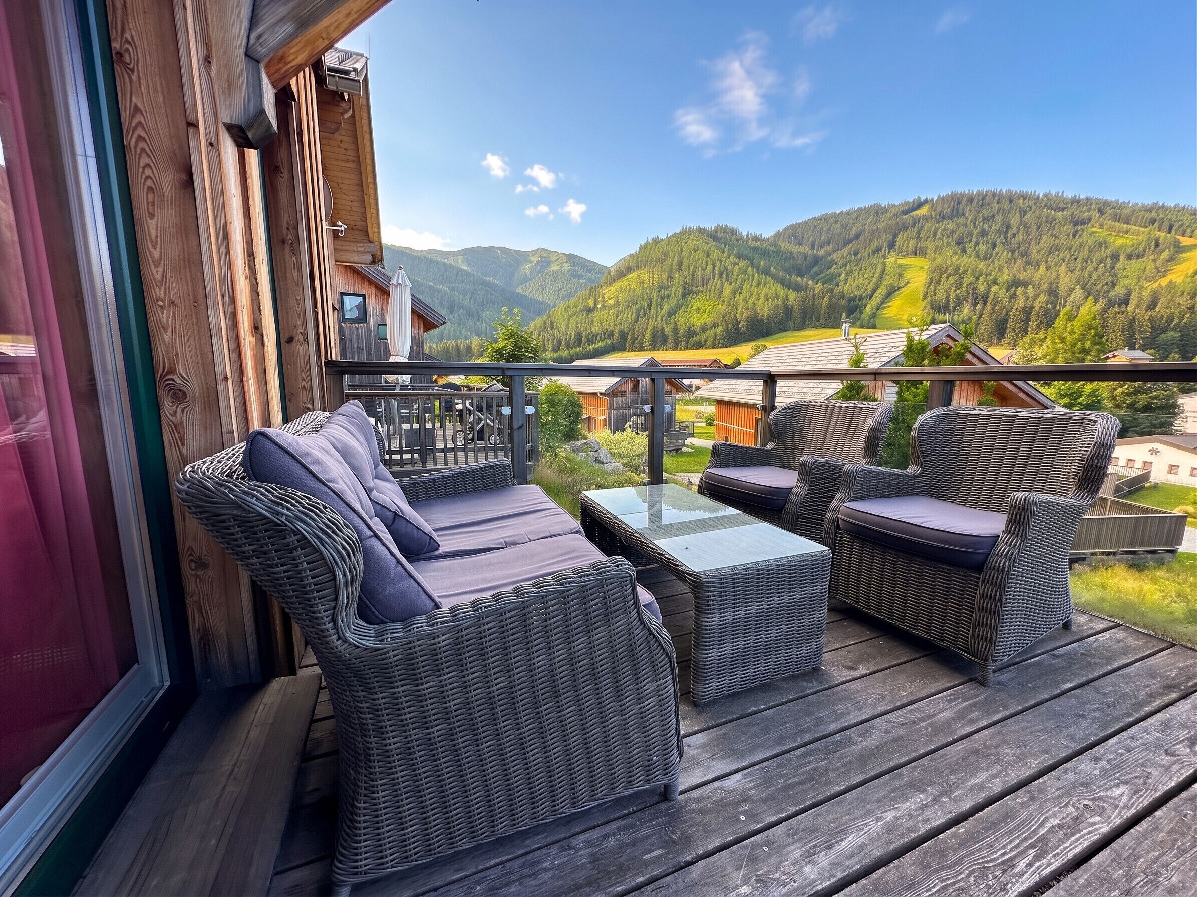 A wooden deck with wicker furniture and a glass table, overlooking a mountainous landscape.