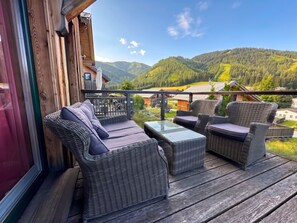 Una terraza de madera con mobiliario de mimbre y una mesa de vidrio, con vistas a un paisaje montañoso.