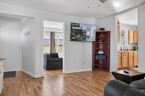 Una sala moderna con un televisor de pantalla plana montado en la pared, un sofá gris, piso de madera y una cocina visible a través de una puerta abierta.