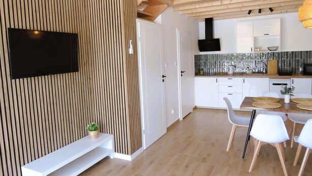 A modern kitchen with a flat-screen TV, wooden paneling, white cabinets, and a dining area with white chairs. - New holiday homes for 6 people, Niechorze (Niechorze)