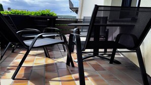 Un patio con sillas y mesa de metal negro, suelo de baldosas y vista a zonas verdes y un edificio al fondo.