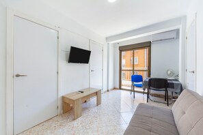 Quarto com sofá, um banco, televisão, escrivaninha com cadeira e uma porta.