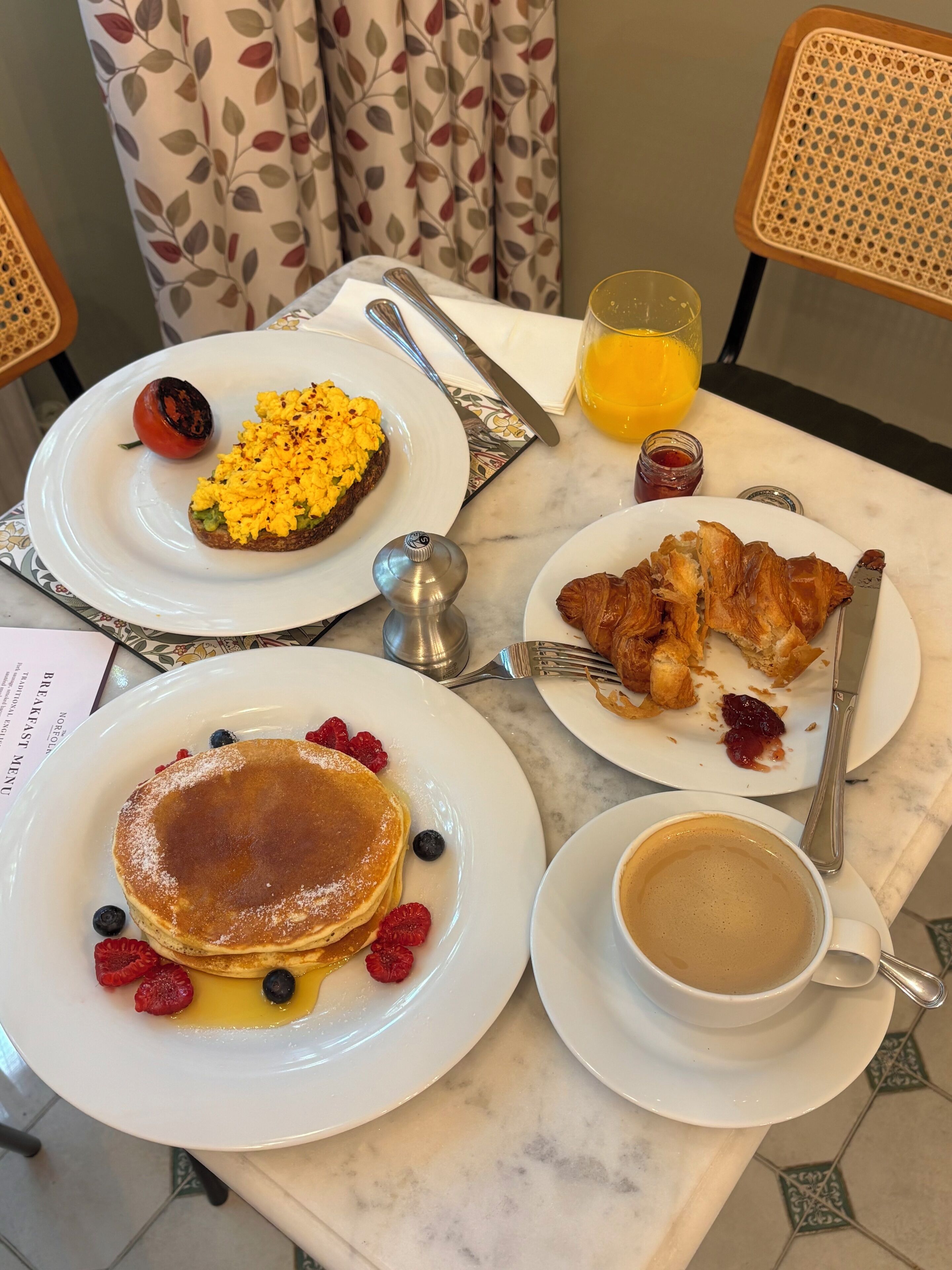 A breakfast table with plates of pancakes, scrambled eggs, croissants, and a cup of coffee.