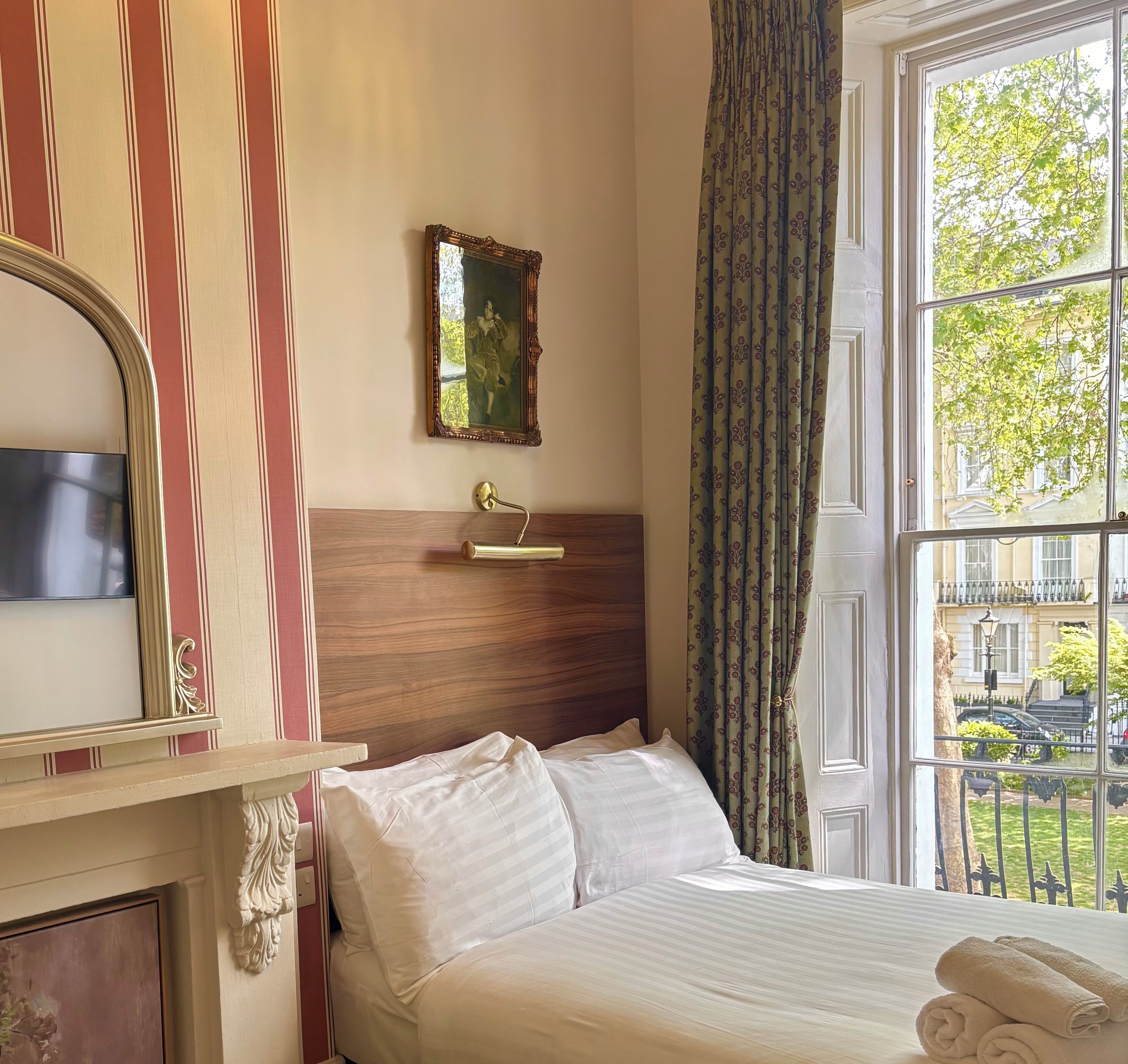 A hotel room with a bed, a mirror, a wall-mounted picture, a window with curtains, and a view of the outside.