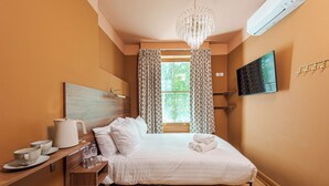 Une chambre à coucher comprenant un lit, une table de chevet avec une bouilloire, des tasses et des bouteilles, un téléviseur fixé au mur et une fenêtre avec des rideaux.