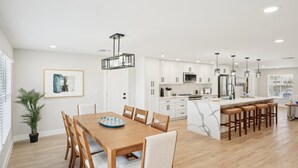 A modern kitchen with white cabinets, a central island with stools, pendant lighting, and a dining area with a wooden table and chairs. - Tranquility Waters (Fort Myers Beach)