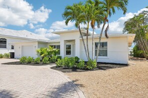 A modern two-story house with a white exterior, a covered patio, and a landscaped garden with palm trees.