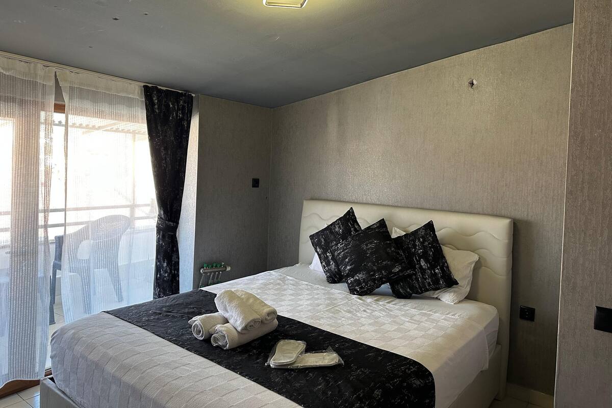 Ein Hotelzimmer mit einem ordentlich bezogenen Bett, einem schwarzen Kopfteil und einem Fenster mit durchsichtigen Vorhängen.