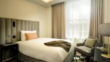 Una habitación de hotel moderna con una cama grande, una mesita de noche con lámpara, una silla y vistas a la ciudad a través de amplios ventanales.