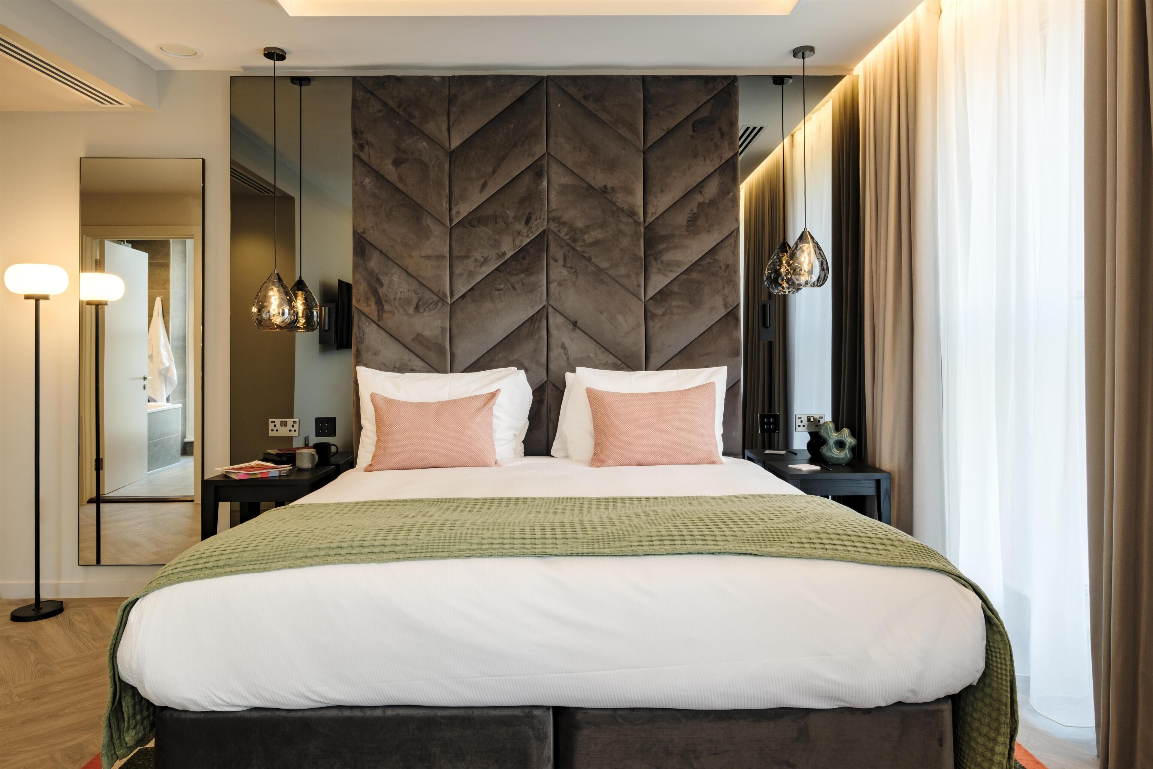 A modern hotel room with a large bed, a mirror, a nightstand, and a green throw blanket.