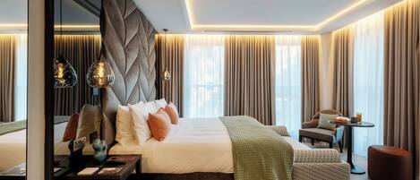 A modern hotel room with a large bed, a bedside table, a small table with a chair, and a large mirror.