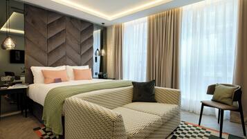 A modern hotel room with a large bed, a sofa, a chair, a desk, and a large window with curtains.
