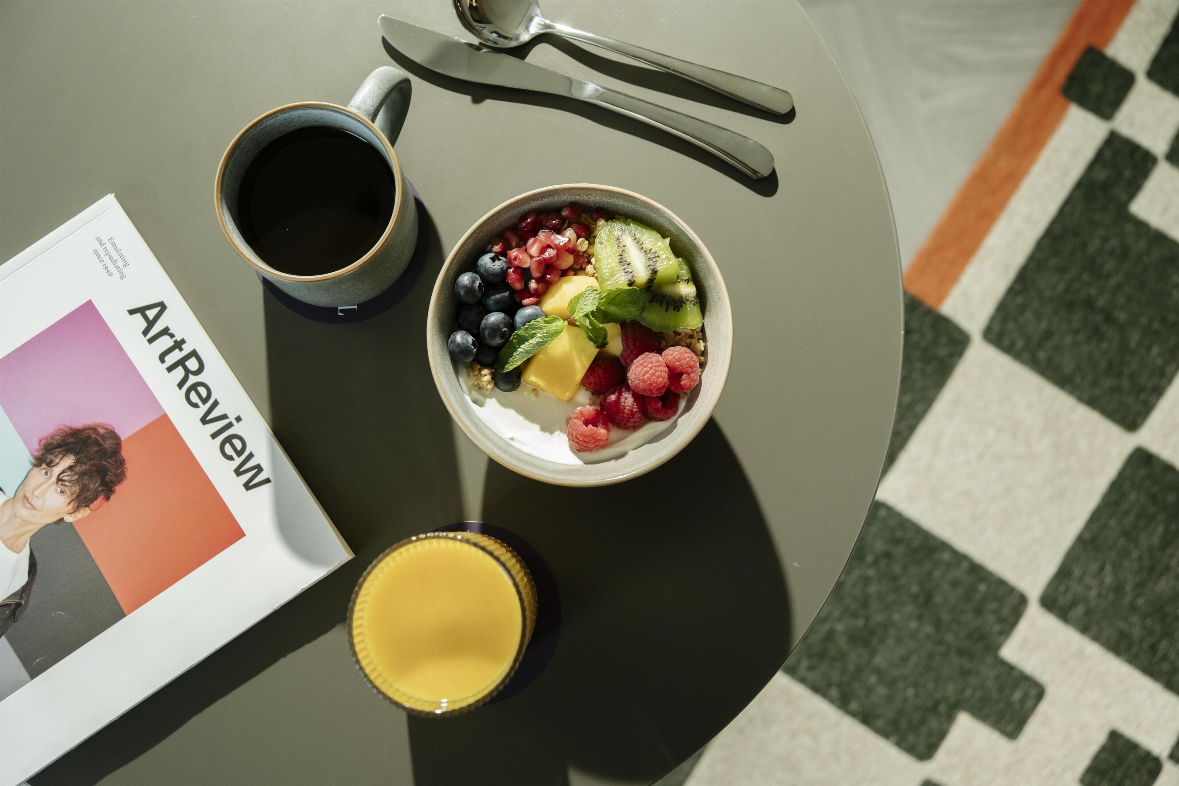 A table with a bowl of fruit, a cup of coffee, a glass of juice, and a magazine.