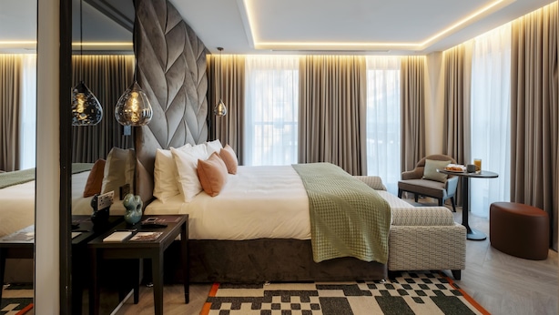 A modern hotel room with a large bed, a desk, a chair, and a small table with a drink.