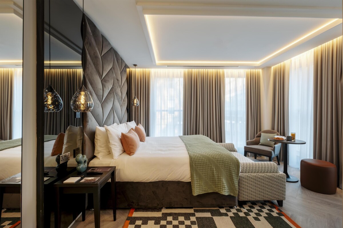 A modern hotel room with a large bed, a desk, a chair, and a small table with a drink.