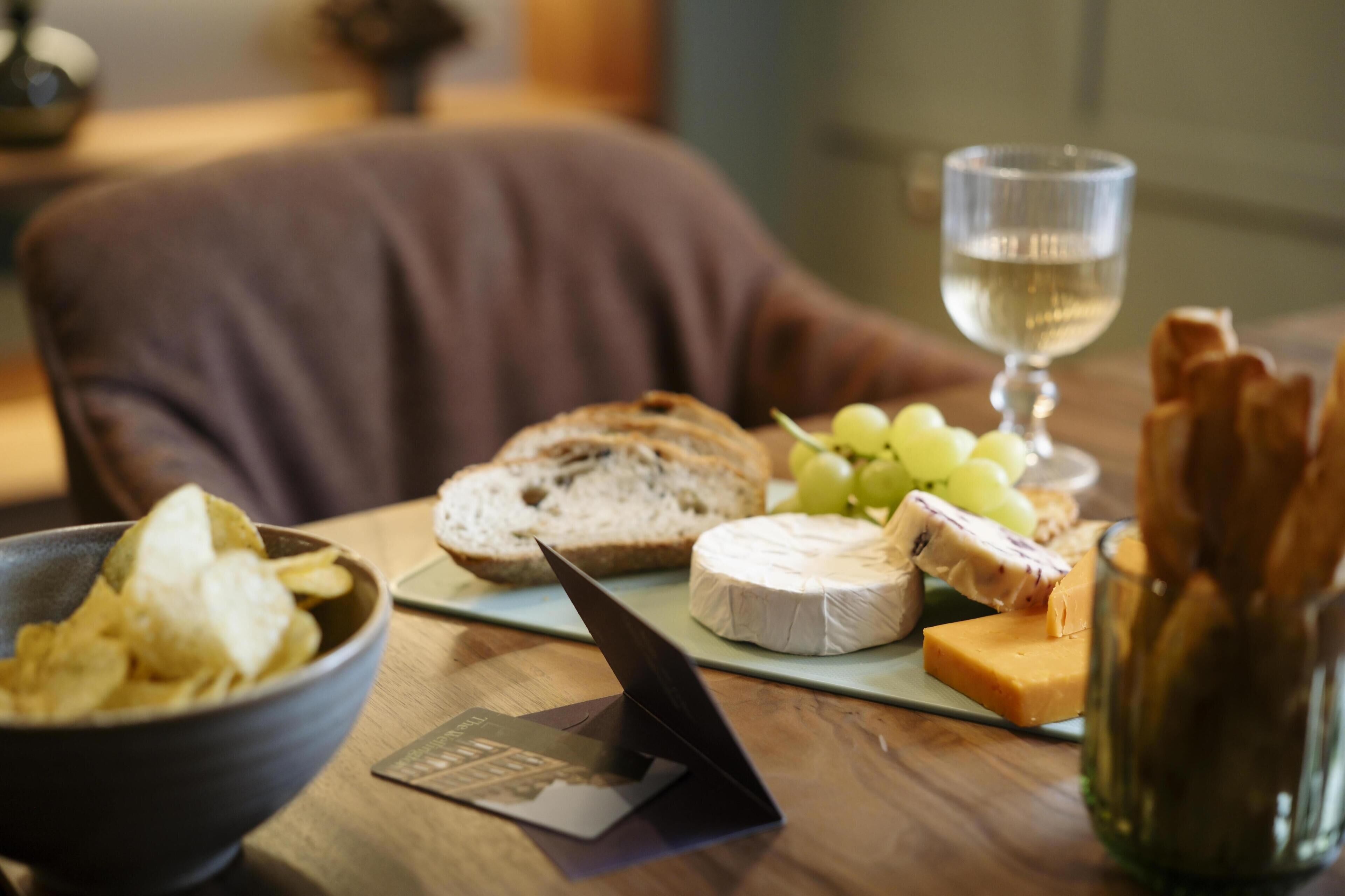 A table with a bowl of chips, a slice of cheese, a piece of bread, a glass of water, and a glass of wine.