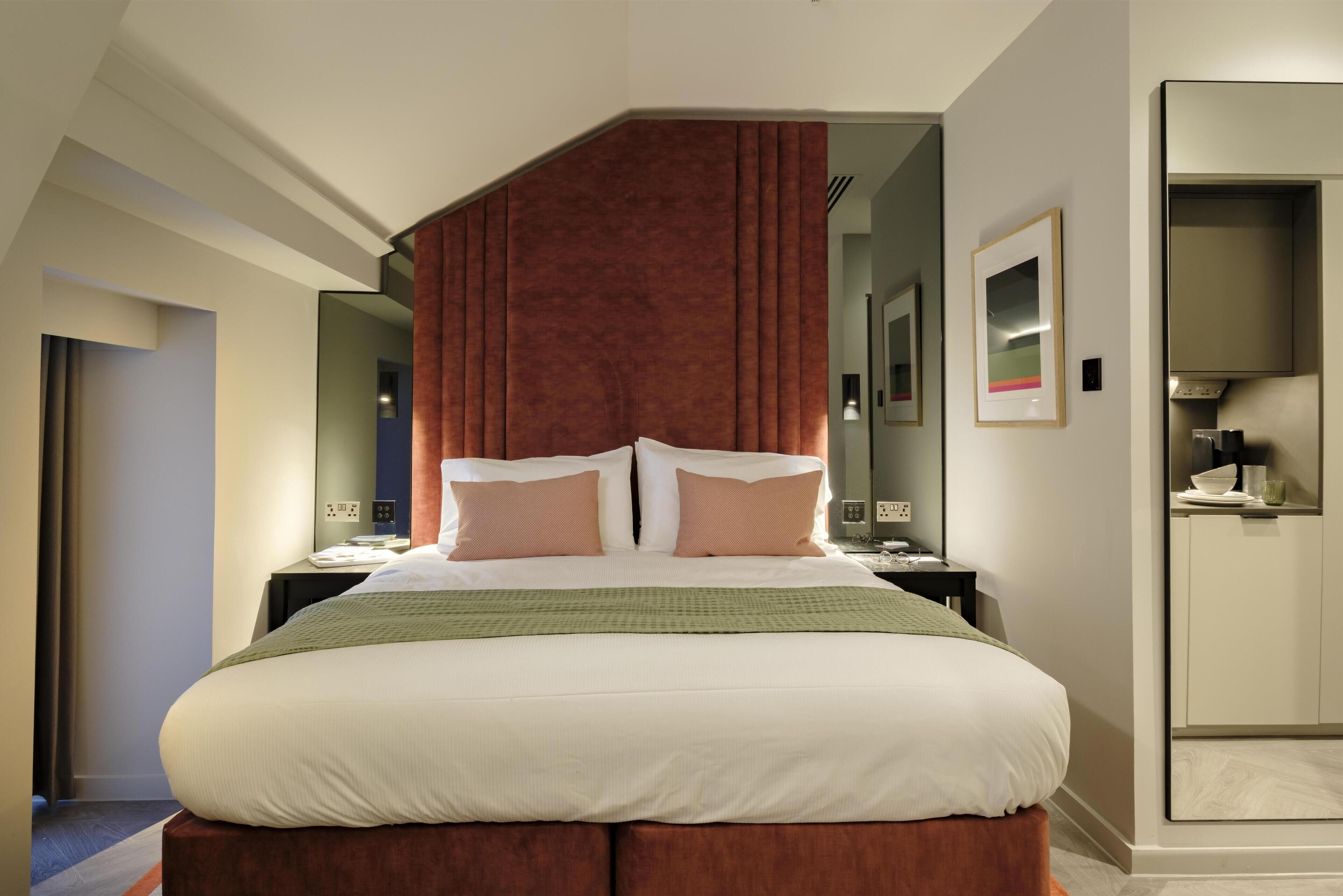 A modern hotel room with a large bed, a wooden headboard, a nightstand, a mirror, and a wardrobe.