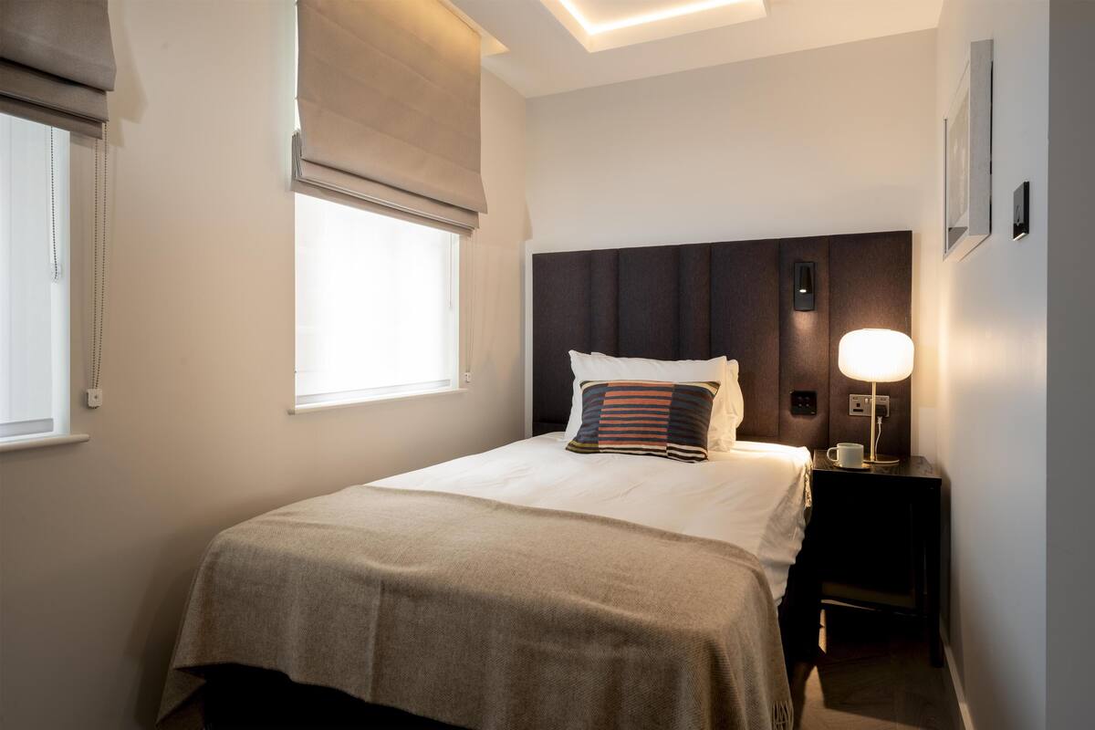 Una cama bien hecha con una colcha color beige, un almohadón a rayas y una lámpara de noche.
