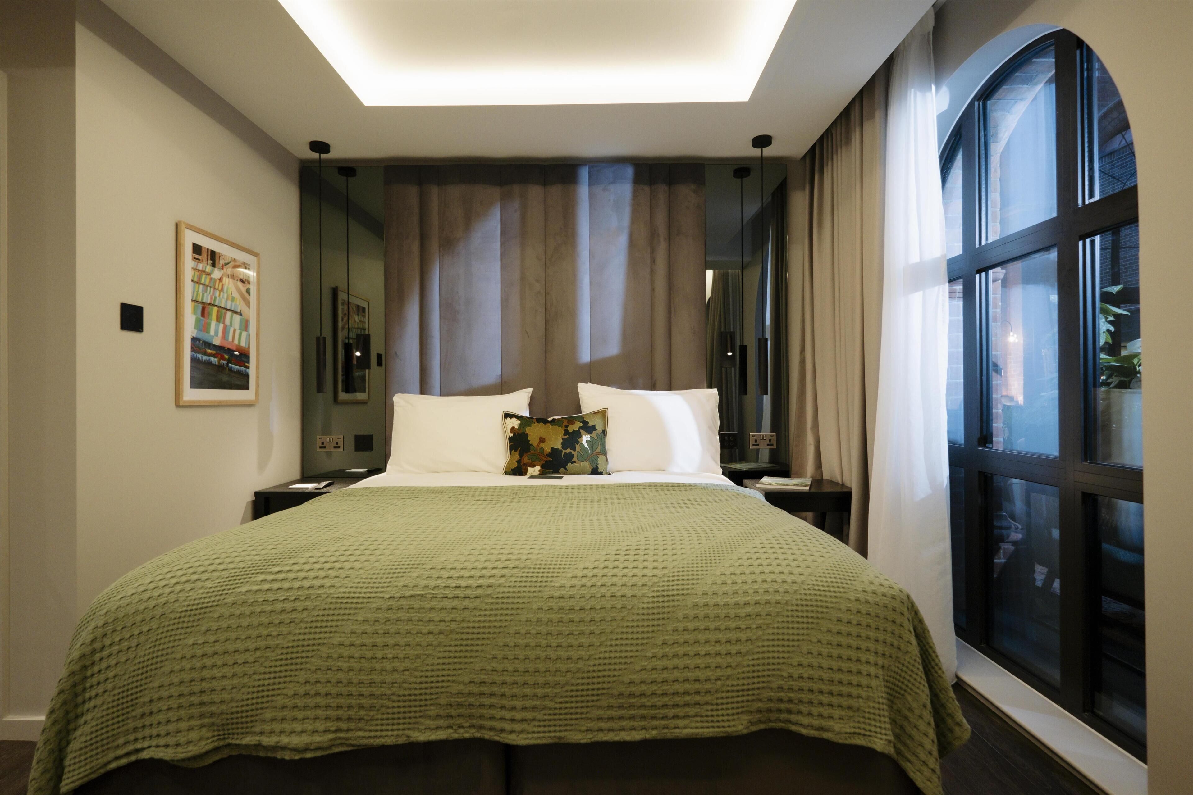Una habitación de hotel moderna con una cama grande, un edredón verde, una mesita de noche, una lámpara, una ventana con cortinas y un cuadro en la pared.
