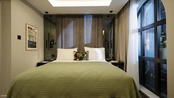 A modern hotel room with a large bed, a green blanket, a nightstand, a lamp, a window with curtains, and a framed picture on the wall.