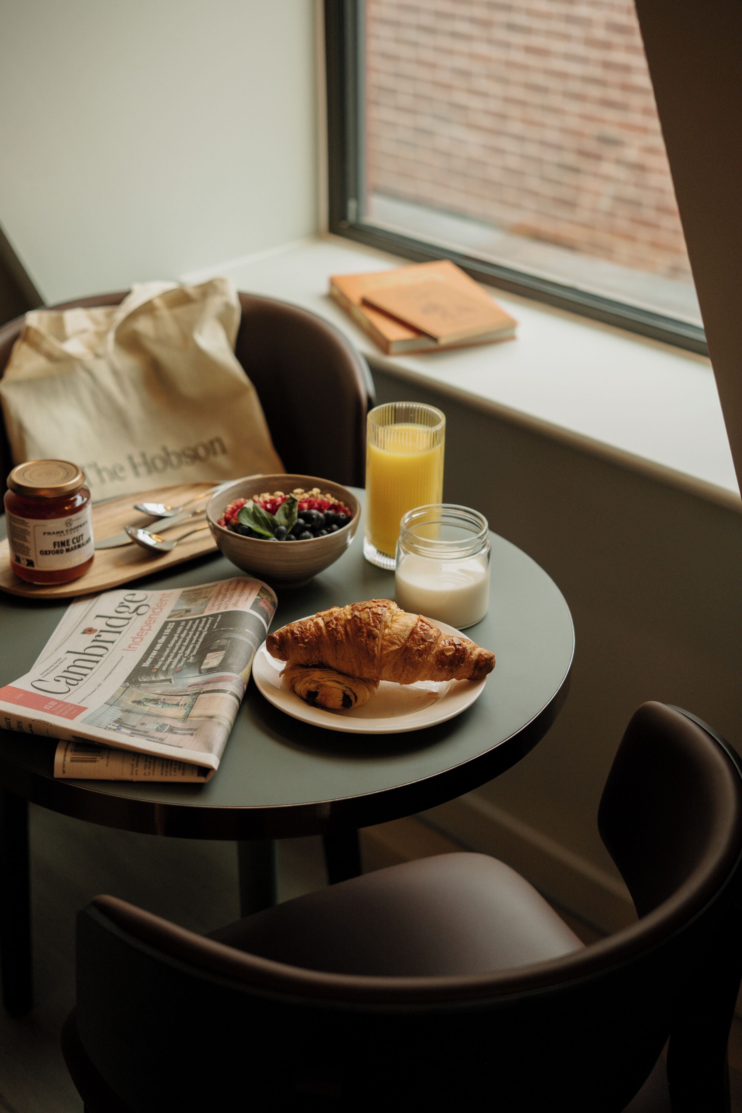 A breakfast setting with a croissant, bowl of fruit, glass of orange juice, milk, and a bag of The Hobson.