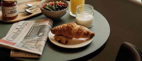 A breakfast setting with a croissant, bowl of fruit, glass of orange juice, milk, and a bag of The Hobson.