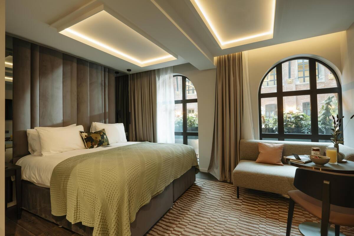 Una habitación de hotel moderna con una cama grande, un sofá, una mesita y vistas a la ciudad a través de amplias ventanas.