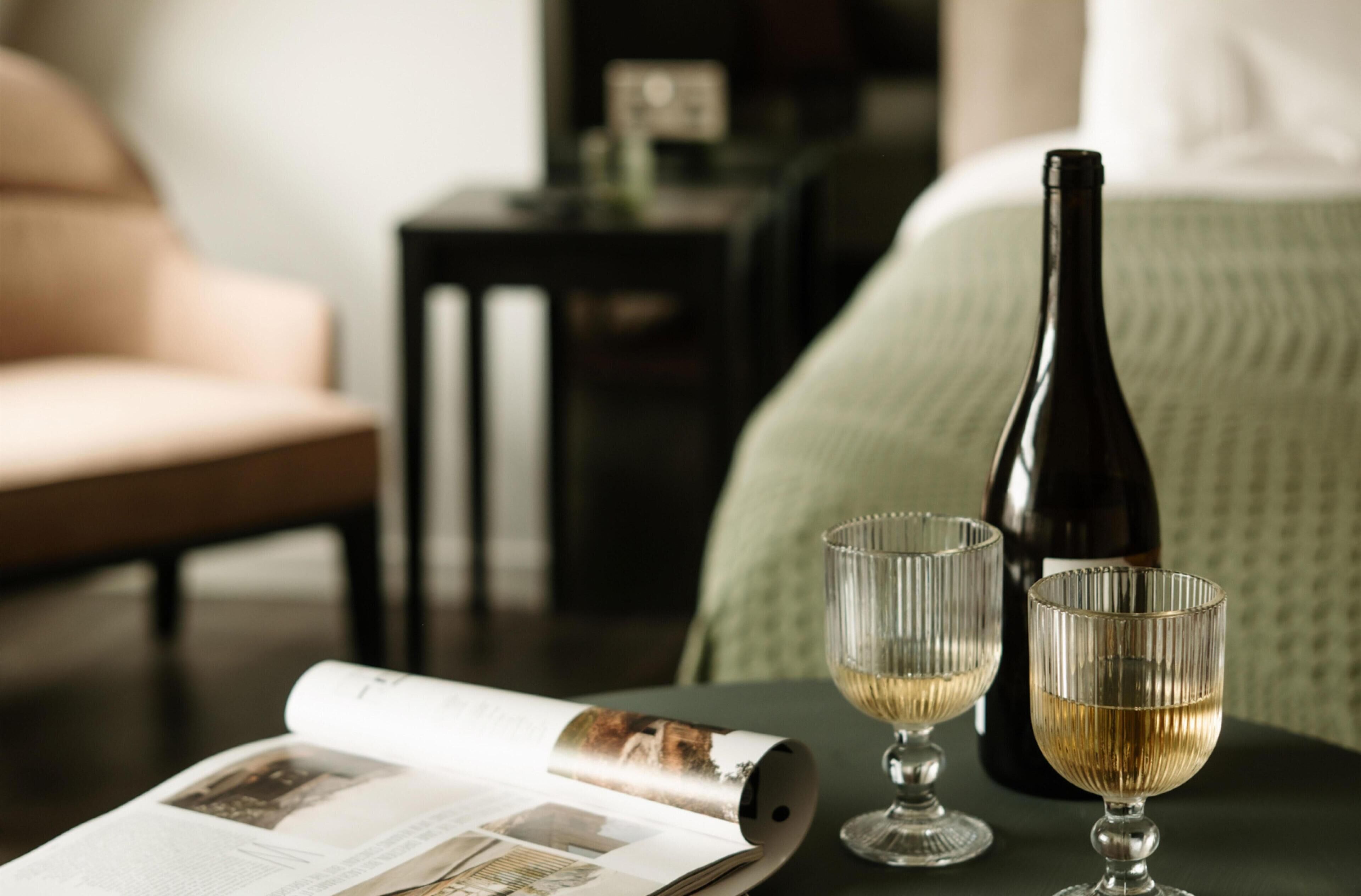 A room with a bed, a table with a magazine, two glasses, and a bottle.