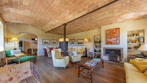 A spacious living room with a brick ceiling, wooden flooring, and a fireplace. There are multiple seating areas with sofas and armchairs, and a coffee table in the center.