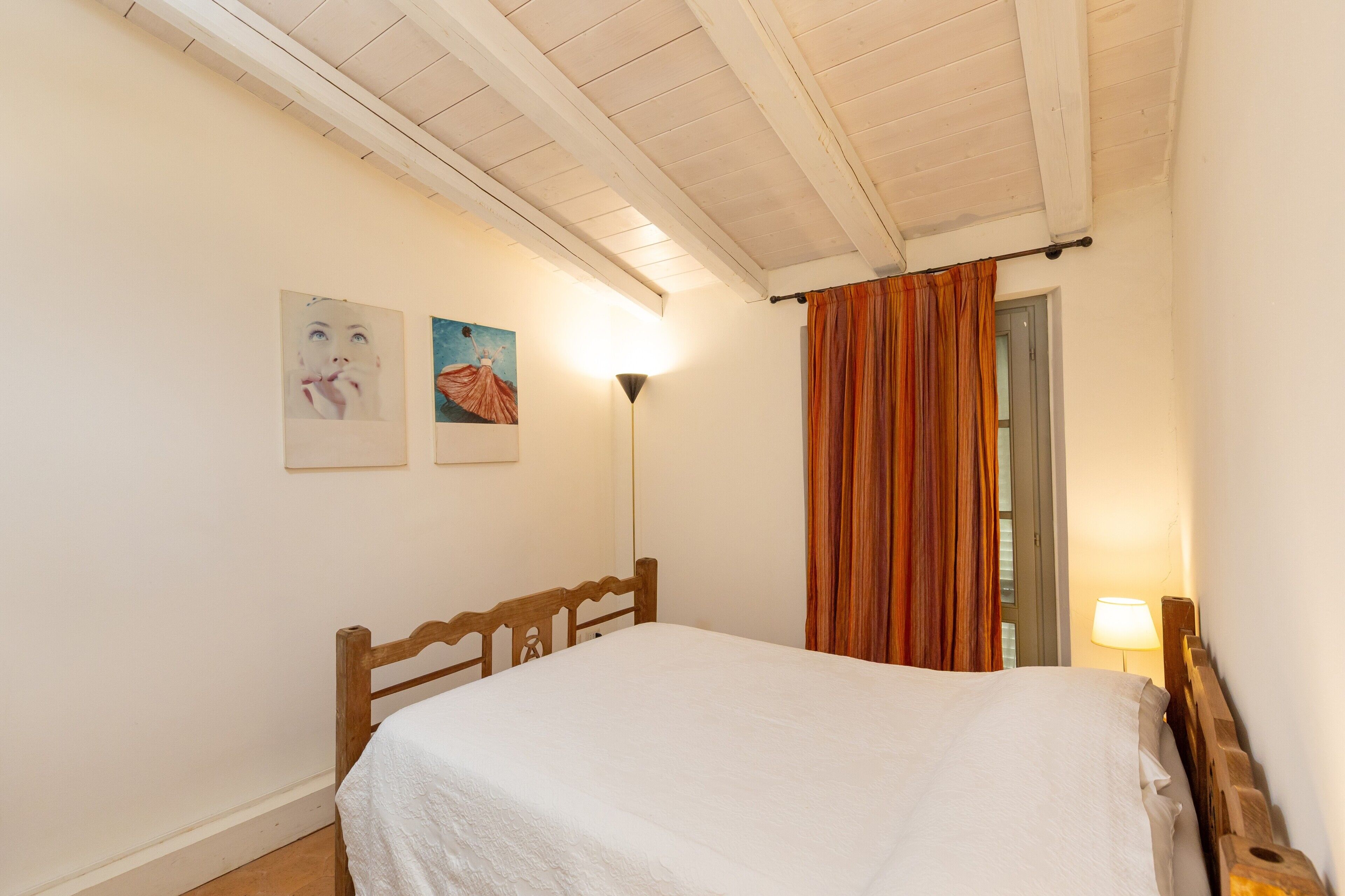 Une chambre à coucher avec un lit en bois, des murs blancs, un plafond avec des poutres apparentes, une lampe sur pied et une fenêtre avec des rideaux.