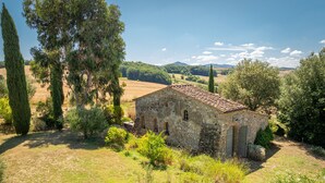 A stone building with a tiled roof surrounded by greenery and fields.