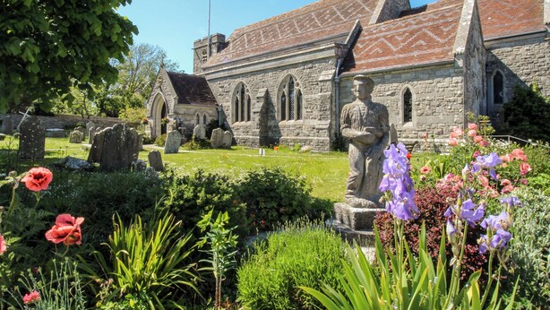 Una chiesa con facciata in pietra, una statua in primo piano e un cimitero con lapidi.