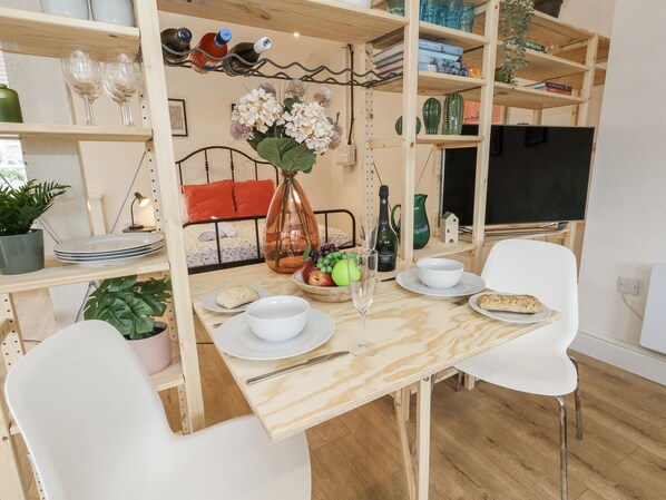 A dining area with a wooden table, white chairs, a vase with flowers, and a bookshelf with various items. - Sea Pink Studio (Swanage)