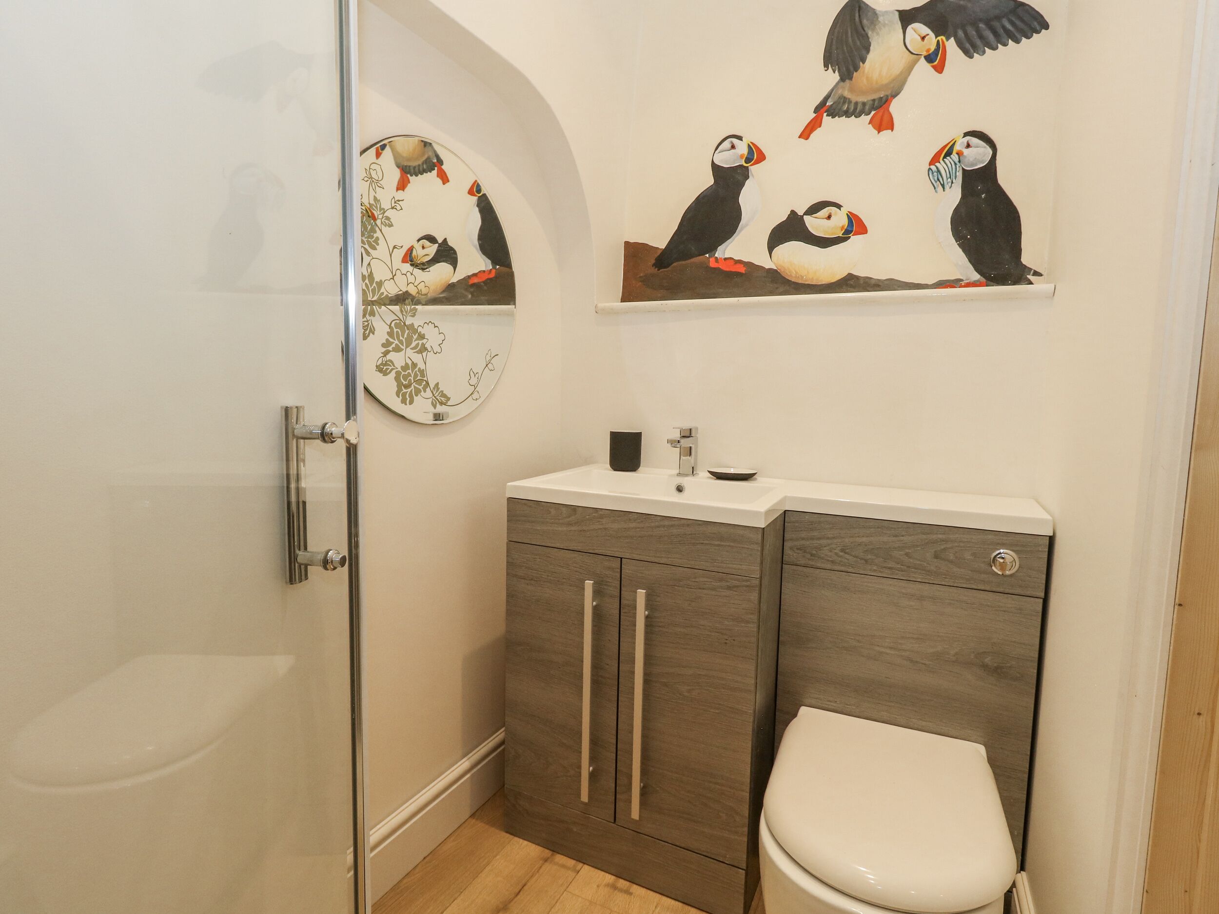 Une salle de bain équipée de toilettes, d’un lavabo et d’une douche. Un meuble mural est fixé au mur, et il porte un tableau représentant des macareux.