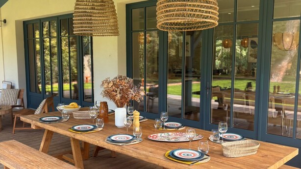 Una terrassa de fusta a l'exterior amb una taula llarga preparada per dinar, amb llums penjants de fibra i vista al jardí a través de grans finestres.