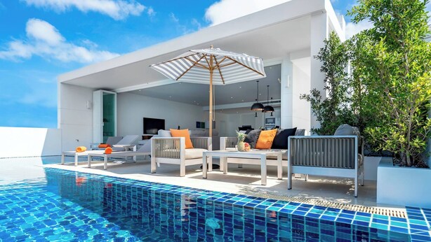 A modern outdoor pool area with lounge chairs and a dining table, surrounded by a white building with large windows.