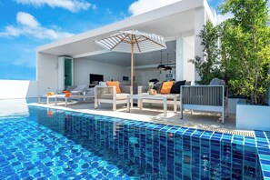 Un área de piscina moderna al aire libre con sillas de descanso y una mesa de comedor, rodeada por un edificio blanco con ventanas grandes.