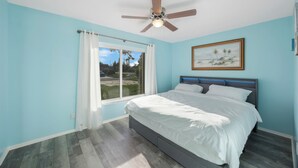 A bedroom with a bed, a ceiling fan, a window with curtains, and a painting on the wall.