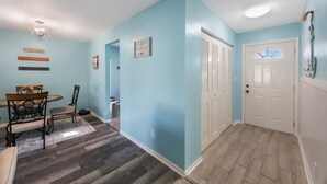 A hallway with light blue walls, a wooden floor, a dining area with a table and chairs, and a white door with a window.
