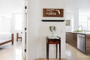 Un intérieur moderne comprenant un lit, une petite table avec un vase, un coin cuisine et une pancarte indiquant « Bienvenue en Floride ».