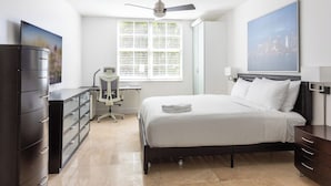 Une chambre à coucher comprenant un lit, un bureau, une chaise, une commode, un ventilateur de plafond et une fenêtre avec des stores.