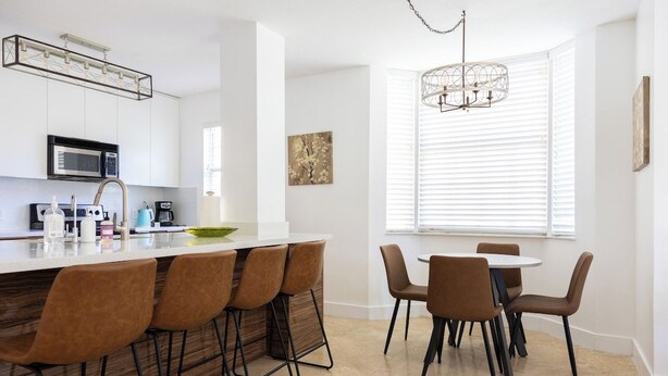 Une cuisine moderne avec un coin bar, une table à manger avec des chaises et une grande fenêtre équipée de stores.