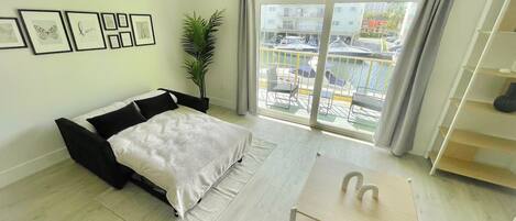 A modern bedroom with a bed, a small table, a potted plant, and a balcony with a view of parked cars and buildings.