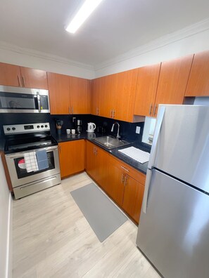 A compact kitchen with wooden cabinets, stainless steel appliances, and a grey rug.