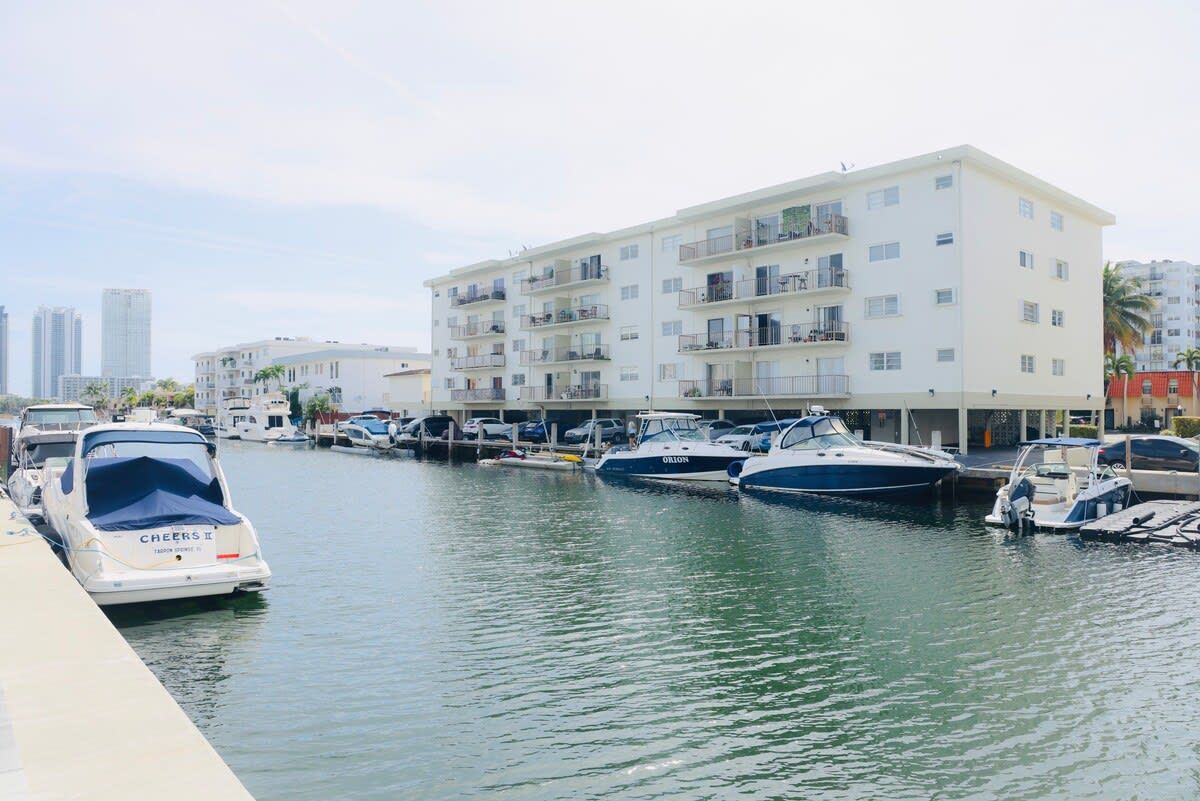 A marina with several boats docked, a white building with balconies, and a clear sky.